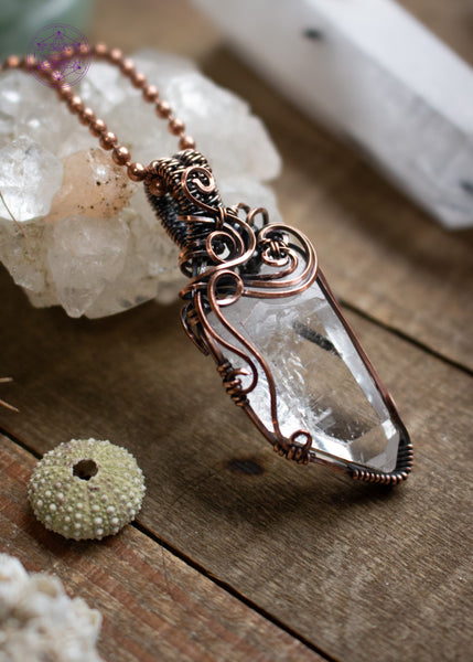 Clear quartz pendant with filigree swirls along the top and side, sitting on a wooden board with crystals and shells around it. The angel shows off the swirls on the left side.