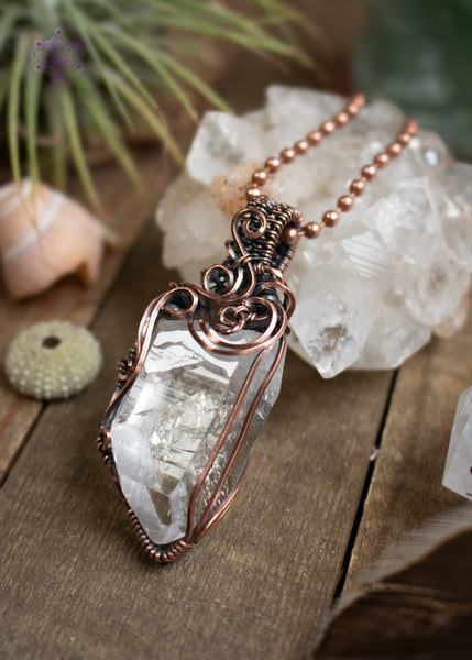 Clear quartz pendant with filigree swirls along the top and side, sitting on a wooden board with crystals and shells around it. The angle shows off the striations along the face of the crystal. 