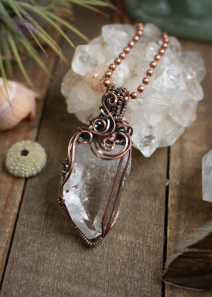 Clear quartz pendant with filigree swirls along the top and side, sitting on a wooden board with crystals and shells around it. The angle shows off the swirls.
