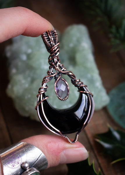 Lilith is a black onyx and faceted amethyst copper wire wrapped pendant. She's a black crescent moon facing upwards (curve side down) and above the moon is the faceted amethyst that captures the light. The bail is elegant and this piece has a very witchy vibe to it. Held in hand to show the light reflections in the crystals.