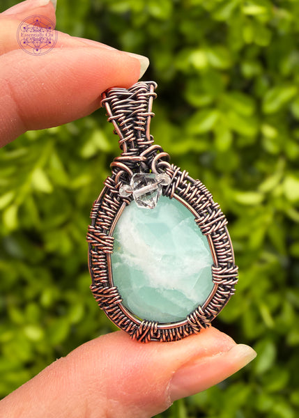 Light mint green Caribbean Calcite crystal handcrafted in copper wire with a woven frame. The bail has an accent stone of diamond quartz and aquamarine to bring in that ocean vibe. Pendant is held in hand with green foliage in the background.