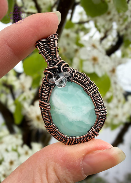 Light green Caribbean Calcite crystal with white inclusions wire wrapped in bare copper wire with a woven frame and oxidized to bring out the details. The bail has accent stones of diamond quartz and aquamarine to bring in that ocean vibe. Shown held with spring white flowers in the background for an elegant look.