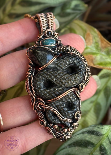 a large gold sheen obsidian snake wrapped in woven copper wire with accent stones of green tournmaline and blue apatite, this one is a statement piece measuring at 3.25" in length. Shown held in hand for size reference.