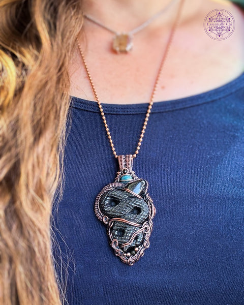 a large gold sheen obsidian snake wrapped in woven copper wire with accent stones of green tournmaline and blue apatite, this one is a statement piece measuring at 3.25" in length. Shown being worn to show how it hangs on the neck.