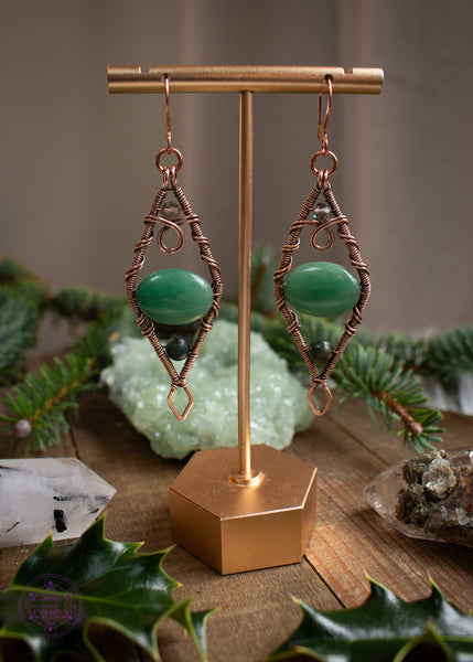 Mossy Spruce Earrings are Copper Wire Wrapped statement earrings in a diamond shape with an oval green aventurine crystal in the middle, moss agate at the bottom point and smoky quartz at the top point. Meant to represent the mossy Sitka Spruce trees in winter with the woodland creatures nestled in it's branches. Shown hanging on an earring display with indoor lighting to show the true color of the stones.
