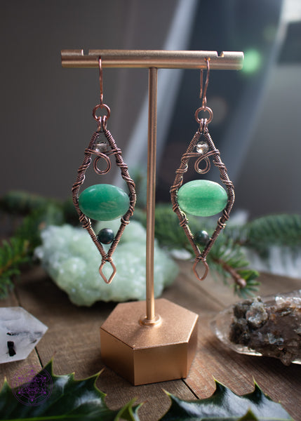 Mossy Spruce Earrings are Copper Wire Wrapped statement earrings in a diamond shape with an oval green aventurine crystal in the middle, moss agate at the bottom point and smoky quartz at the top point. Meant to represent the mossy Sitka Spruce trees in winter with the woodland creatures nestled in it's branches. Shown hanging on an earring display with the light illuminating them from behind.