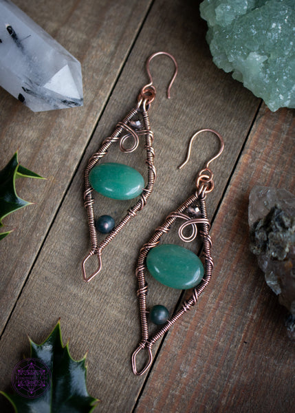 Mossy Spruce Earrings are Copper Wire Wrapped statement earrings in a diamond shape with an oval green aventurine crystal in the middle, moss agate at the bottom point and smoky quartz at the top point. Meant to represent the mossy Sitka Spruce trees in winter with the woodland creatures nestled in it's branches. Shown on a wooden table laying flat with crystals and holly leaves around it.