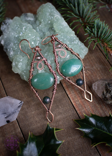 Mossy Spruce Earrings are Copper Wire Wrapped statement earrings in a diamond shape with an oval green aventurine crystal in the middle, moss agate at the bottom point and smoky quartz at the top point. Meant to represent the mossy Sitka Spruce trees in winter with the woodland creatures nestled in it's branches. Shown laying on a prehnite crystal with holly leaves and spruce branches around it on a wooden table.