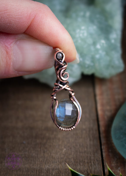Copper wire wrapped faceted Labradorite pendant with a small Labradorite bead in the bail. This is a dainty filigree design meant to resemble a rain drop. Held in hand with a green prehnite crystal behind on a wooden board.