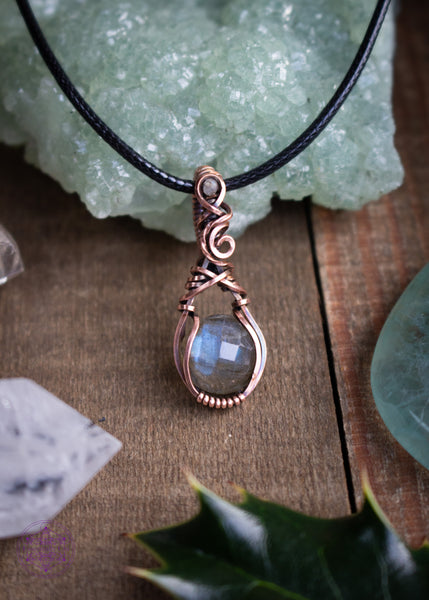 Copper wire wrapped faceted Labradorite pendant with a small Labradorite bead in the bail. This is a dainty filigree design meant to resemble a rain drop. Shown leaning against a green prehnite crystal on a wooden board with crystals and holly leaves around it for a winter feel.