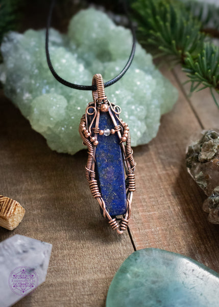 Lapis Lazuli copper wire wrapped necklace with a moonstone accent bead. The design looks like the curves of a woman holding a small moonstone bead. The Lapiz Lazuli is so dark with the flecks of pyrite within that mimmic the night sky. Hence why I named this pendant Asteria after the goddess of the night sky. Shown sitting against a green prehnite crystal on a wooden table with spruce branches and crystals around it.