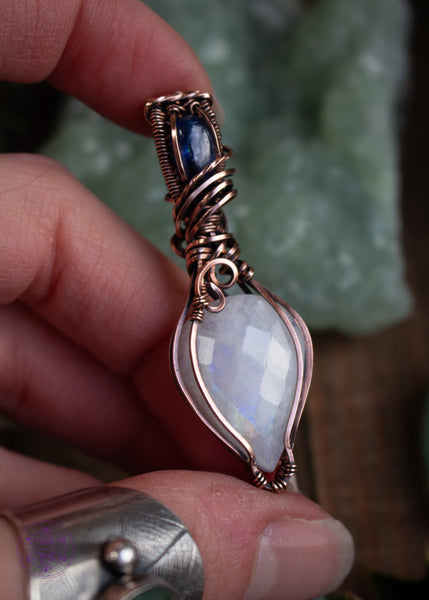 This Rainbow Moonstone and Blue Kyanite necklace is wrapped in copper wire with a teardrop moonstone pointing down, reflecting a rainbow flash off the facets of the stone. The bail has a brilliant dark blue kyanite and swirls around the bail like winter winds whirling around the snow. This pendant reminds me of a fresh blanket of snow on a cloudless winter night.  Shown in hand to bring out the rainbow in the moonstone reflecting off the light.