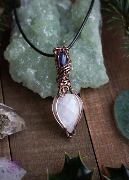 This Rainbow Moonstone and Blue Kyanite necklace is wrapped in copper wire with a teardrop moonstone pointing down, reflecting a rainbow flash off the facets of the stone. The bail has a brilliant dark blue kyanite and swirls around the bail like winter winds whirling around the snow. This pendant reminds me of a fresh blanket of snow on a cloudless winter night.  Shown on a wooden board with green crystals, holly leaves, and spruce twigs to set the christmas feeling.