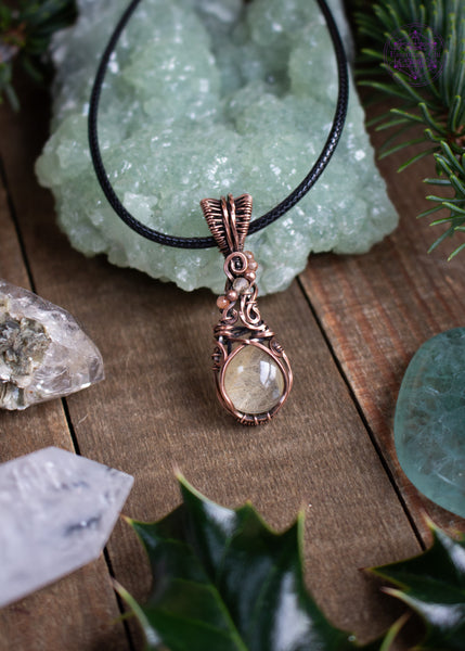 This copper wire wrapped pendant has a small round golden rutile crystal and accent crystal beads of sunstone and citrine that catch the light and really make this pendant sparkle like stardust! Picture shows the pendant laying against a green prehnite crystal with crystals, spruce twigs and holly leaves around it. Also shown is the black cotton waxed cord it comes with.