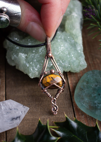 Tiger Eye copper wire wrapped pendant with a geometric design of an elongated diamond with a faceted golden tiger eye crystal and citrine and copper beads over the tiger eye. simple and elegant. Shown on a table with crystals, holly leaves, and spruce needles around it.