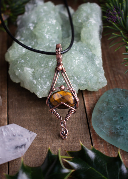 Tiger Eye copper wire wrapped pendant with a geometric design of an elongated diamond with a faceted golden tiger eye crystal and citrine and copper beads over the tiger eye. simple and elegant. Shown on a table with crystals, holly leaves, and spruce needles around it.
