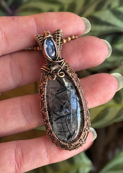 an oval cut black tourmaline quartz crystal wrapped with copper wire like a basket weave and a blue kyanite and smoky quartz accent crystals in the bail. Held in hand to show size.