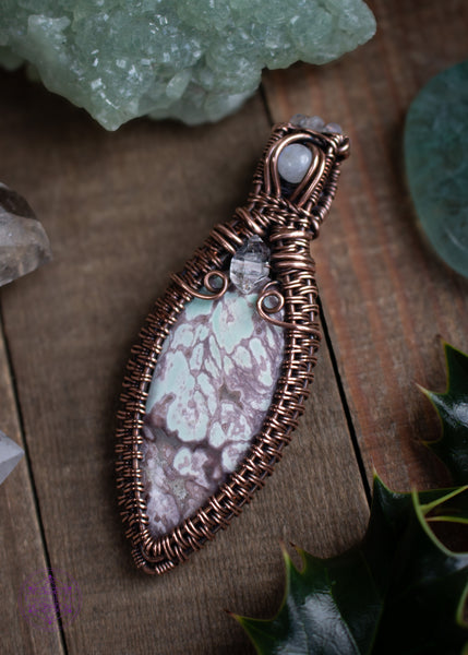 Copper wire wrapped Variscite pendant with accent stones of diamond quartz and moonstone made to look like a winter goddess, named Chione after the greek goddess of snow. Shown on a wooden table with spruce branches around it. The variscite has a beautiful minty green color with veins of grey throughout it.