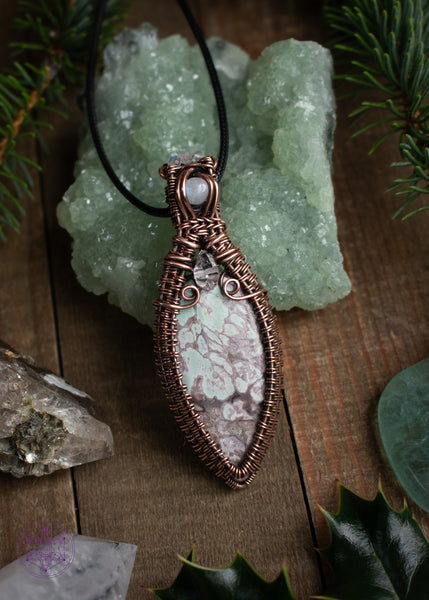 Copper wire wrapped Variscite pendant with accent stones of diamond quartz and moonstone made to look like a winter goddess, named Chione after the greek goddess of snow. Shown leaning against a prehnite crystal on a wooden table with spruce branches around it.