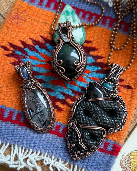 A combination of all Transformation pendants available. The Black Tourmaline quartz necklace, the Rainbow Obsidian Sphere Pendant, and the Gold Sheen obsidian snake pendant. Sitting on a mini navajo rug with colors of orange and lavender. 