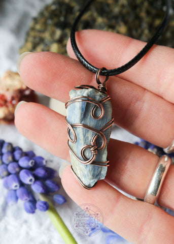 This copper wire wrapped pendant shown in hand for size reference has 4 swirls along the face of the blue kyanite and is connecting the two middle swirls together. This raw crystal has a triangular shape where its flat on the back and pointed in the middle and 1.75" long. Raw Blue Kyanite Wire Wrapped Necklace for energy protection, featuring a simple, everyday wear design that enhances communication, alignment, tranquility, restful sleep, and dream recall.