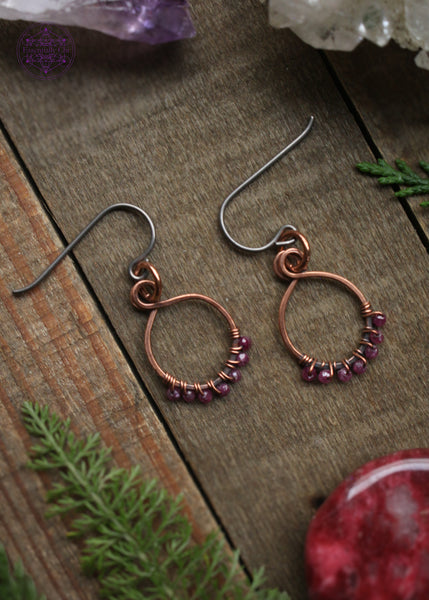 Pair of dainty copper hoops with small faceted rubys woven along the bottom of them. Sitting on a wooden table with crystals and foliage around them.