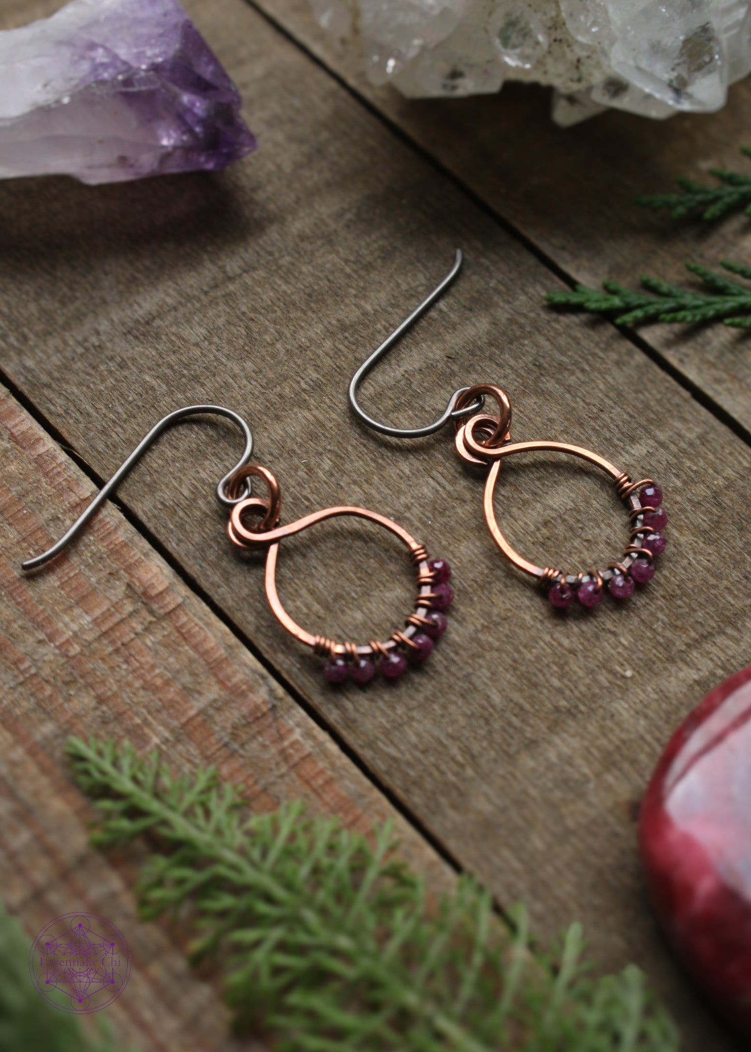 Pair of dainty copper hoops with small faceted rubys woven along the bottom of them. Sitting on a wooden table with the light shining on the copper so perfectly and crystals and foliage around them.
