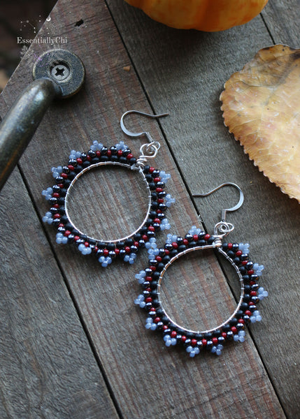 Dark Witch Beaded Hoop Earrings featuring a matte black, pomegranate red, hematite, and chalcedony-colored seed bead pattern. Suspended from sterling silver ear hooks, these 2.5-inch earrings exude enchanting allure