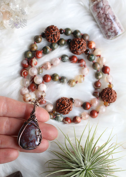 Longing for the serenity of a forest walk? Let the Amanita Forest Mala carry you away. Hand-knotted with Flower Agate, Brecciated Red Jasper, Rainforest Jasper, Smoky Quartz, and 4 Rudraksha seeds, this 72-bead mala necklace is designed to help nature lovers, energy healers, and nature witches ground themselves and open their hearts to new opportunities. Crafted with a copper wire-wrapped Red Moss Agate pendant, this unique piece is the perfect companion for your meditation practice. Beaded Mala
