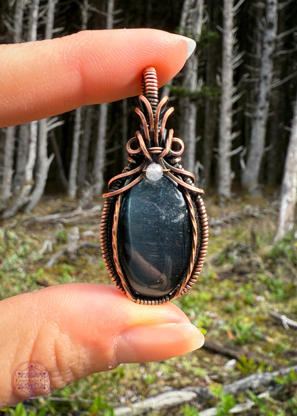 Beautiful Blue Tigers eye pendant with a bail shaped like a pineapple like top and a dark topaz crystal holding them together. The front of the pendant has two twisted wires going up the side. Simple and elegant. The background is a mossy forest floor with the skeletons of the shore pine tree for dramatic effect.