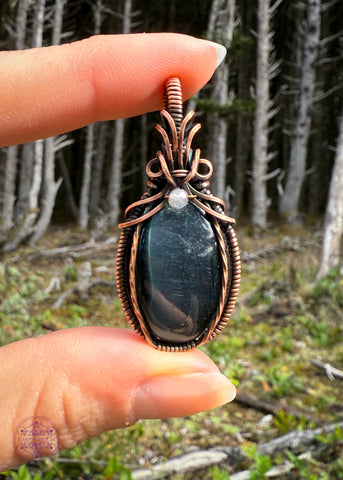 Beautiful Blue Tigers eye pendant with a bail shaped like a pineapple like top and a dark topaz crystal holding them together. The front of the pendant has two twisted wires going up the side. Simple and elegant. The background is a mossy forest floor with the skeletons of the shore pine tree for dramatic effect.