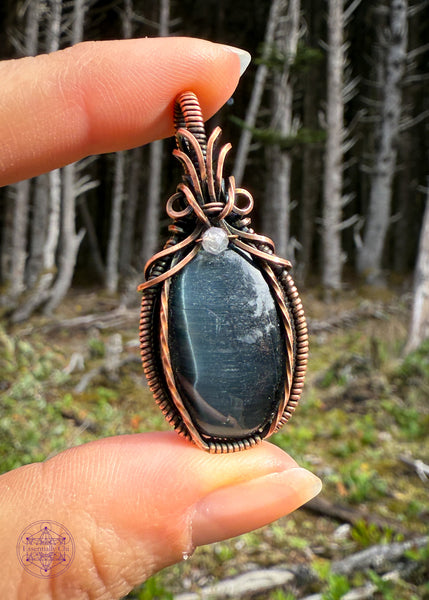 Beautiful Blue Tigers eye pendant with a bail shaped like a pineapple like top and a dark topaz crystal holding them together. The front of the pendant has two twisted wires going up the side. Held in fingers at a side angle to catch the light. Simple and elegant. The background is a mossy forest floor with the skeletons of the shore pine tree for dramatic effect.