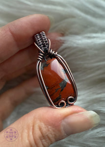 Red jasper copper wire wrapped pendant with grey quartz marbling throughout the stone. The wrap just hugs the outside of the rectangular stone to show all of the beautiful red jasper. The bail is a tight weave with one swoop on each side for the simple elegant look. Held in hand with a white faux fur background.