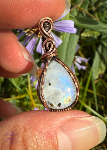 Beautiful Rainforest moonstone teardrop pendant wrapped in copper wire is a dainty small crystal pendant. The front square wires are twisted over the edge of the crystal and the bail has a few elegant swoops. Held in hand in front of purple asters to enhance the elegant look.