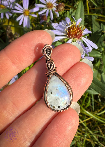 Beautiful Rainforest moonstone teardrop pendant wrapped in copper wire is a dainty small crystal pendant. The front square wires are twisted over the edge of the crystal and the bail has a few elegant swoops. Pendant shown on fingers to show size reference. (Two finger widths long).
