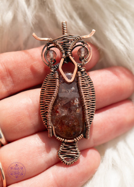 copper wire wrapped great horned owl pendant with a fire quartz belly, wire woven wings, and smoky quartz eyes held in hand with a white faux fur background.
