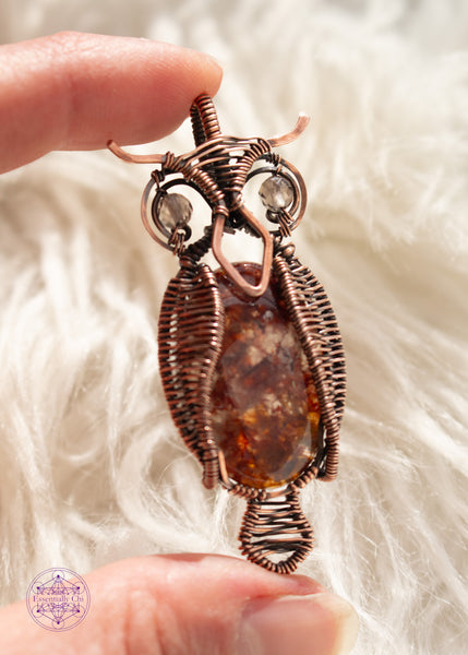 copper wire wrapped great horned owl pendant with a fire quartz belly, wire woven wings, and smoky quartz eyes held in hand with a white faux fur background.