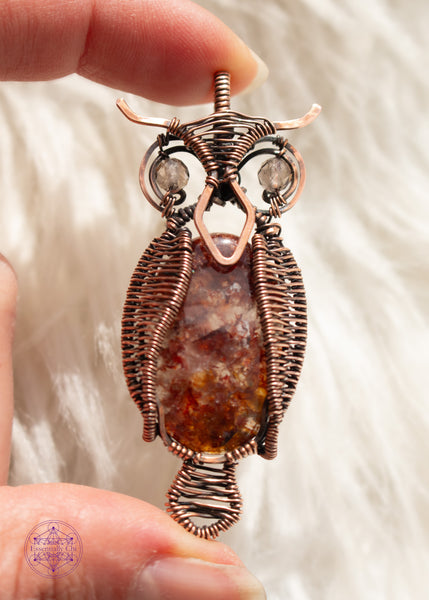 copper wire wrapped great horned owl pendant with a fire quartz belly, wire woven wings, and smoky quartz eyes held in hand with a white faux fur background.