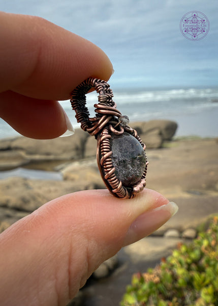 mini garden quartz pendant held in fingers the sides and the ocean and beach in the background. The bail is small and thick weave with a small smoky quartz at the base of bail and three small copper balls at the base of the pendant to add extra security to the frame. The garden quartz crystal is a 10mm circle wrapped in copper wire.