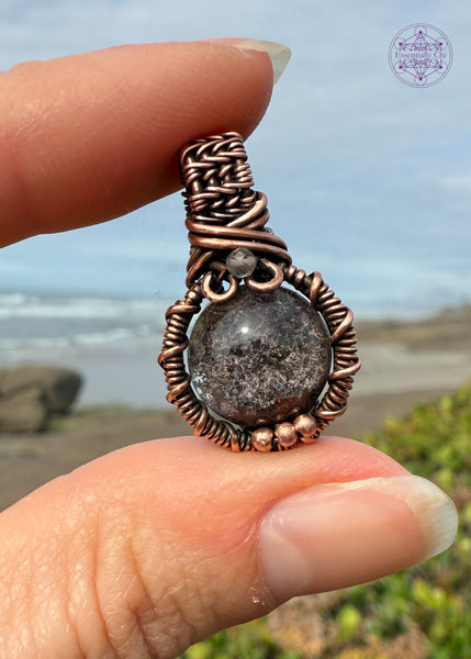 mini garden quartz pendant held in fingers with the ocean and beach behind it to show off the colors in the garden quartz as well as the reflect that this pendant reminds me of a barnacle or some sea creature. The bail is small and thick weave with a small smoky quartz at the base of bail and three small copper balls at the base of the pendant to add extra security to the frame. 