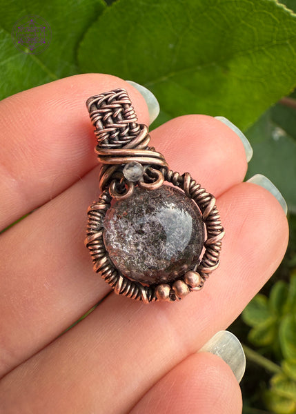 mini garden quartz pendant held in fingers to show size with green leaves in the background. The bail is small and thick weave with a small smoky quartz at the base of bail and three small copper balls at the base of the pendant to add extra security to the frame. The garden quartz crystal is a 10mm circle wrapped in copper wire.