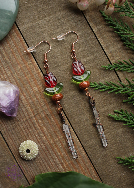 Pair of long dangle earrings on a wooden table. The Earrings hang from rose gold plated hooks and have acrylic red tulip beads, then goldstone and copper wire wrapped quartz points dangling. Foliage and crystals around it.
