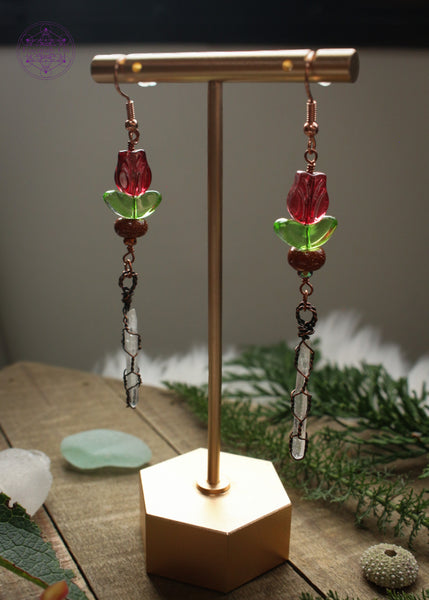 Pair of long dangle earrings on a earring stand. The Earrings hang from rose gold plated hooks and have acrylic red tulip beads, then goldstone and copper wire wrapped quartz points dangling. Foliage and sea glass around it on a wooden base.