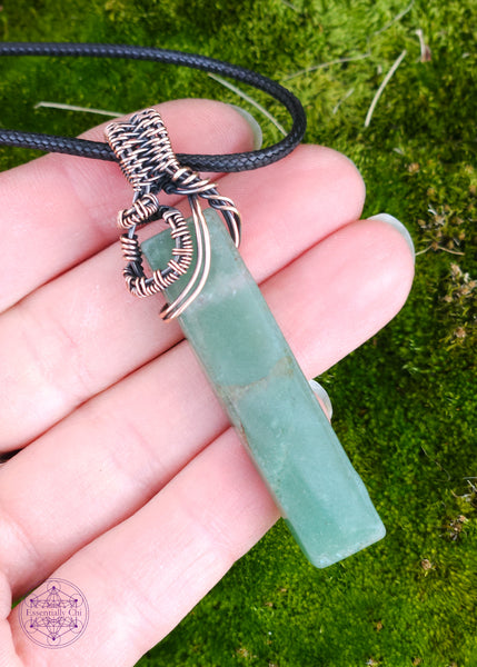 green aventurine pendant wrapped with copper wire and a black cotton waxed cord. The crystal is a rectangle and the copper wrapping is close to the bail showing more of the stone. Shown in hand for size reference. 