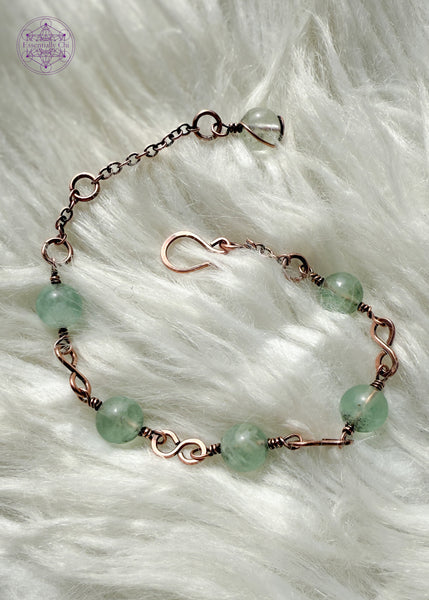 Copper wire wrapped bracelet features 8mm green Fluorite beads individually wrapped with figure 8 Infinity links in-between for an elegant bracelet. Showing the whole bracelet on a faux white fur blanket to see hook clasp and beaded extension chain and green fluorite charm.
