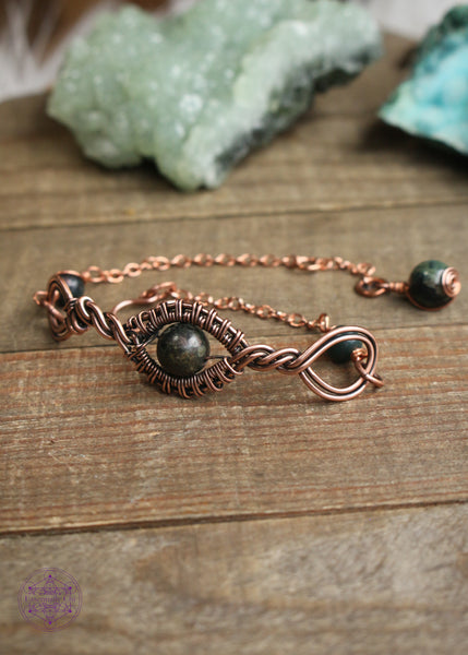 Copper wire wrapped bracelet features a Kambaba Jasper bead wrapped in twisted copper design with loops on each end, with accent connectors on the sides with small moss agate beads and a charm at the end of kambaba jasper and a spiral design. Shown sitting on a wooden boad and green crystals in the background.