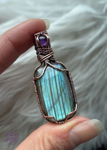 gorgeous blue flash labradorite with dark vertical streaks through it is wrapped in copper wire and held at the corners so it didn't cover the flashy parts. The bail is a woven pattern and a small dark purple amethyst stone to add to the depths of this pendant. 