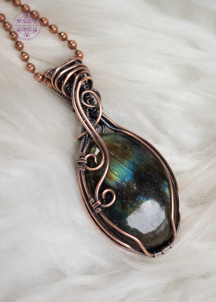 Multi-flash labradorite copper wire wrapped necklace with a simple design of a few swirls going down the left side of the crystal and a flowing bail. In-between the flashes are deep darkness, like peering into the ocean with the reflections in the darkness. Shown on a white faux fur background.