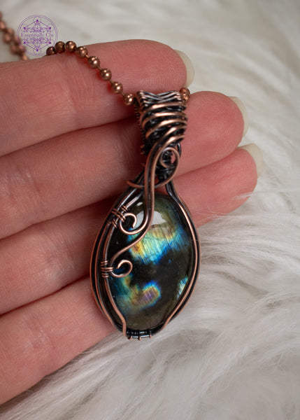 Multi-flash labradorite copper wire wrapped necklace with a simple design of a few swirls going down the left side of the crystal and a flowing bail. In between the flashes are deep darkness, like peering into the ocean with the reflections in the darkness. Held in hand over a white faux fur background.