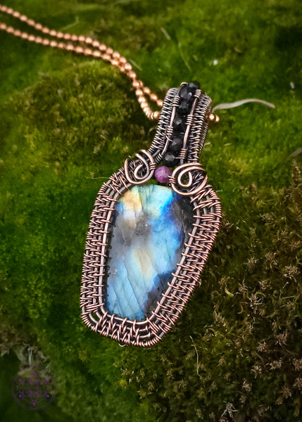 Copper wire wrapped labradorite coffin pendant with a red tourmaline accent stone at the top of the coffin to symbolize blood color, and then black spinel beads along the center of the bail. nice thick wrap style to create the ultimate statement piece. Labradorite has mainly blue flash with a few dark spots and yellow to orange colors. on a mossy rock for contrast.